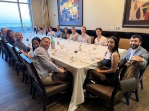 A group of people sit around a large U-shaped table at a restaurant, smiling at the camera during an Attorney Retreat. There are drinks and table settings, with a scenic window view and colorful artwork in the background, capturing the spirit of team building at Ansbacher Law.