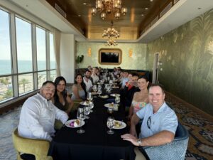 A group of people dressed in semi-formal attire sit around a long table in a bright, elegant dining room with large windows overlooking the ocean, enjoying a meal together during an Ansbacher Law attorney retreat focused on team building.