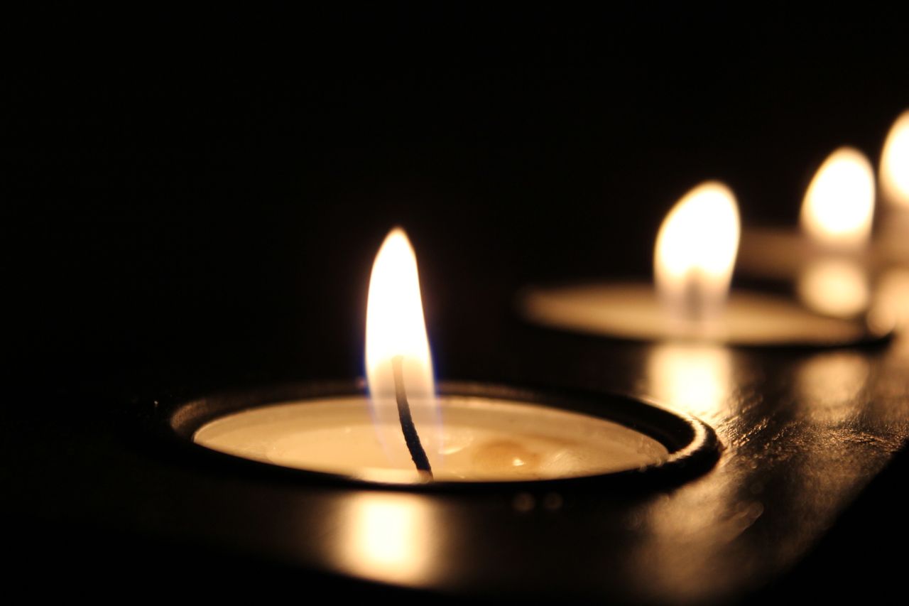 A close-up of a lit tealight candle with a soft, warm flame, set in a dark environment. Blurred flames from additional candles are visible in the background, creating a peaceful and calm atmosphere.